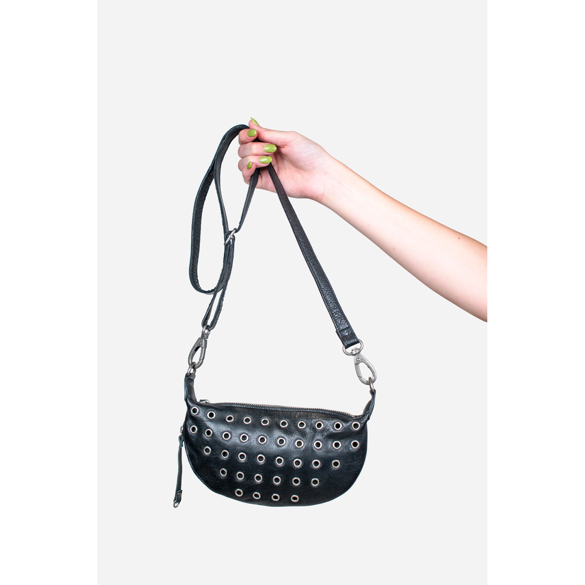 A black faux-leather crescent-shaped shoulder bag with silver metal grommets, held by a hand against a light gray background.